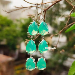 Peut inclure: Boucles d'oreilles pendantes argentées avec trois niveaux de pierres précieuses en forme de goutte d'eau, de couleur teal. Les boucles d'oreilles sont suspendues à un crochet et portent le texte "KSBJewels".