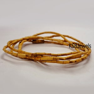 Pu&ograve; includere: Un primo piano di una collana di perline di legno color marrone chiaro. La collana &egrave; composta da piccole perline rettangolari che sono infilate insieme per formare un lungo filo. La collana &egrave; arrotolata su una superficie bianca.