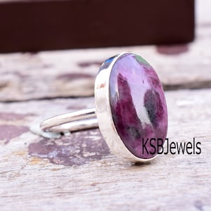 May include: A silver ring featuring a pink and purple gemstone. The ring is set in a silver band with a smooth, polished finish. The oval-shaped gemstone has a distinctive swirling pattern. The ring is displayed on a wooden surface. The text 'KSBJewels' is printed on the surface.