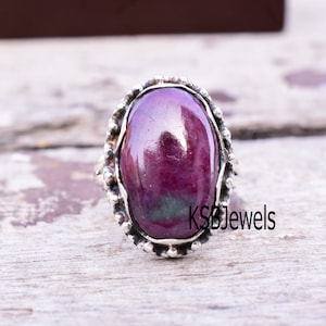 Puede incluir: Un anillo de plata con una gema grande, ovalada y de color rojo intenso. La gema pulida está engastada en una banda de plata texturizada y ornamentada. El texto "KSB Jewels" es visible en el anillo.