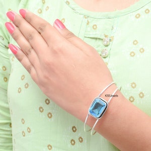 May include: A silver bangle bracelet with a large, light blue gemstone set in a silver bezel. The bracelet is open and has a simple design. The text "KSBJewels" is visible on the bracelet.