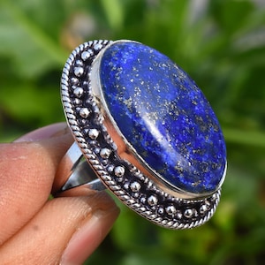 Op de afbeelding: Een zilveren ring met een grote ovale lapis lazuli-edelsteen. De steen is diepblauw met gouden vlekken. De ring heeft een decoratieve band met kleine kralen.