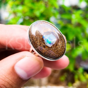 May include: Oval-shaped ring featuring a large, iridescent opal stone set in a silver band. The opal displays a mix of colors, including blue, green, and gold, set against a dark brown matrix. The ring is held by a hand against a blurred green background.