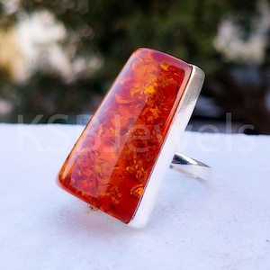 Puede incluir: Un anillo rectangular de ámbar con una banda de plata. La piedra de ámbar tiene un color rojo-naranja intenso con patrones internos. El anillo está engastado en un engaste de plata pulida.