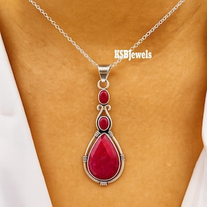 May include: A silver pendant necklace with a large, teardrop-shaped, red gemstone. Two smaller red gemstones are above the larger stone. The pendant hangs from a delicate silver chain. The text "KSBjewels" is visible.