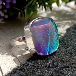 Puede incluir: Un anillo de plata con una piedra preciosa iridiscente de forma cuadrada. La piedra preciosa presenta una combinación de colores púrpura, azul y verde con una superficie texturizada.