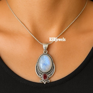 May include: A silver pendant necklace featuring an oval moonstone and a small red gemstone. The pendant has an ornate silver setting with a beaded border and decorative swirls. The necklace is displayed against a dark background, with the text "KSBjewels" visible.