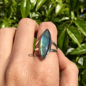 Puede incluir: Un anillo de plata con una gran piedra preciosa de labradorita verde iridiscente engastada en forma de marquesa. El anillo está en un dedo, con follaje verde en el fondo.