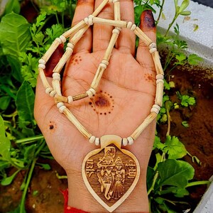 Pu&ograve; includere: Una collana di legno con un ciondolo a forma di cuore che presenta un'immagine intagliata di una divinit&agrave;. La collana &egrave; realizzata con perline di legno naturale e ha un colore marrone chiaro.