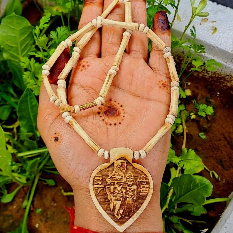 Pu&ograve; includere: Una collana di legno con un ciondolo a forma di cuore che presenta un'immagine intagliata di una divinit&agrave;. La collana &egrave; realizzata con perline di legno naturale e ha un colore marrone chiaro.