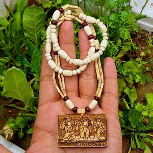 Pu&ograve; includere: Una collana di legno con un ciondolo che presenta un'immagine intagliata di quattro figure. Il ciondolo &egrave; marrone e ha il testo "Shri Ram Darshabar" inciso sul fondo. La collana &egrave; fatta di perline bianche e marroni.