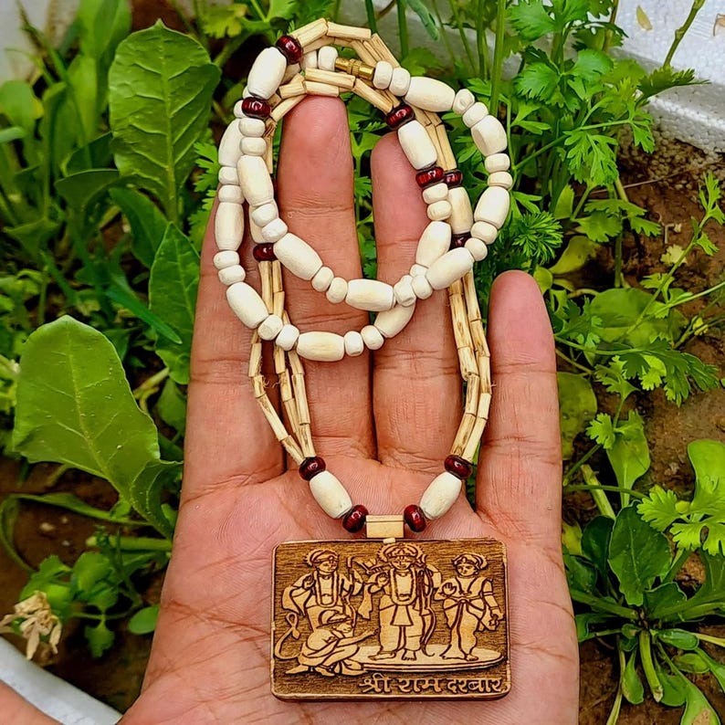 Pu&ograve; includere: Una collana di legno con un ciondolo che presenta un'immagine intagliata di quattro figure. Il ciondolo &egrave; marrone e ha il testo "Shri Ram Darshabar" inciso sul fondo. La collana &egrave; fatta di perline bianche e marroni.