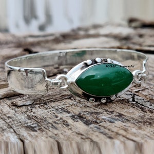 May include: A silver bangle bracelet with a green oval gemstone set in a silver bezel. The bracelet has a hinged opening and is decorated with small silver beads. The text "KSB_Jewels" is visible on the bracelet.