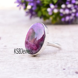 May include: A silver ring featuring a pink and green oval gemstone. The ring is displayed on a wooden surface.