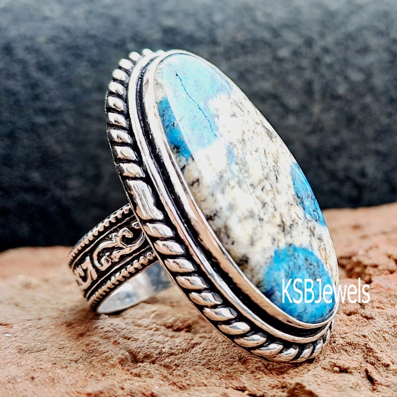 May include: A silver ring featuring a blue and white oval stone set in a rope-like design. The ring is on a brown background. KSBJewels is printed on the stone.