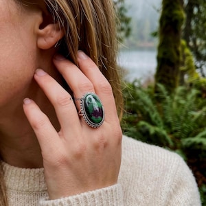 Puede incluir: Un anillo de plata con una gran piedra cabujón de forma ovalada con un patrón de color verde oscuro y magenta. La piedra está engastada en un bisel de metal plateado con diseño de cuerda trenzada. El texto KJBJewels es visible en la piedra.