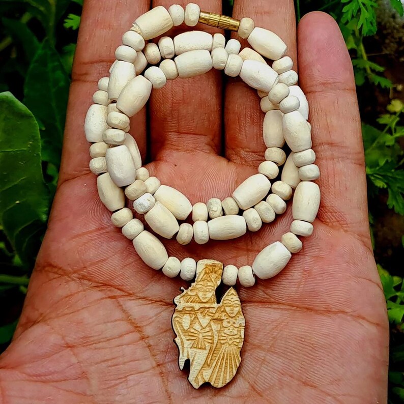 Pu&ograve; includere: Una collana di perline di legno bianco con un ciondolo in legno intagliato con tre figure. Il ciondolo &egrave; di colore marrone chiaro.