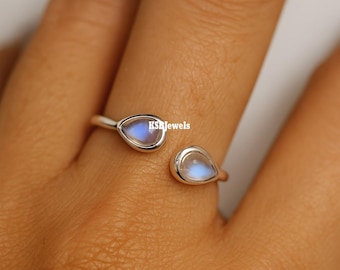 May include: Open-style silver ring featuring two pear-shaped moonstone cabochons. The moonstones have a blue and white iridescent glow. The ring is set against a neutral background. The text "KSJewels" is visible on the ring.
