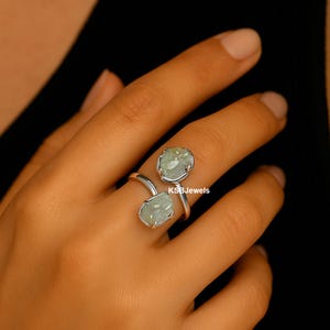 Op de afbeelding: Een zilveren ring met twee ovale, lichtgroene edelstenen. Het ontwerp van de ring wikkelt zich om de vinger, met de edelstenen in een bezel-zetting. De merknaam "KSBJewels" is zichtbaar.