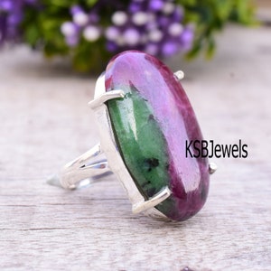 Puede incluir: Un anillo de plata con una gran piedra ovalada verde y rosa. El anillo está engastado con garras y tiene un acabado pulido. KSBJewels está impreso en la superficie de madera.