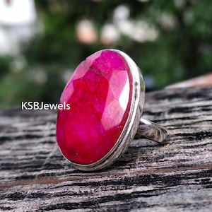 Op de afbeelding: Een zilveren ring met een grote, ovaal geslepen, gefacetteerde robijn. De robijn heeft een levendige rode kleur. De ring ligt op een houten oppervlak met textuur. De tekst "KSBJewels" is zichtbaar.
