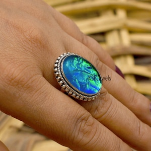 May include: A silver ring featuring a large oval blue and green opal gemstone set in a beaded bezel. The ring is worn on a finger.