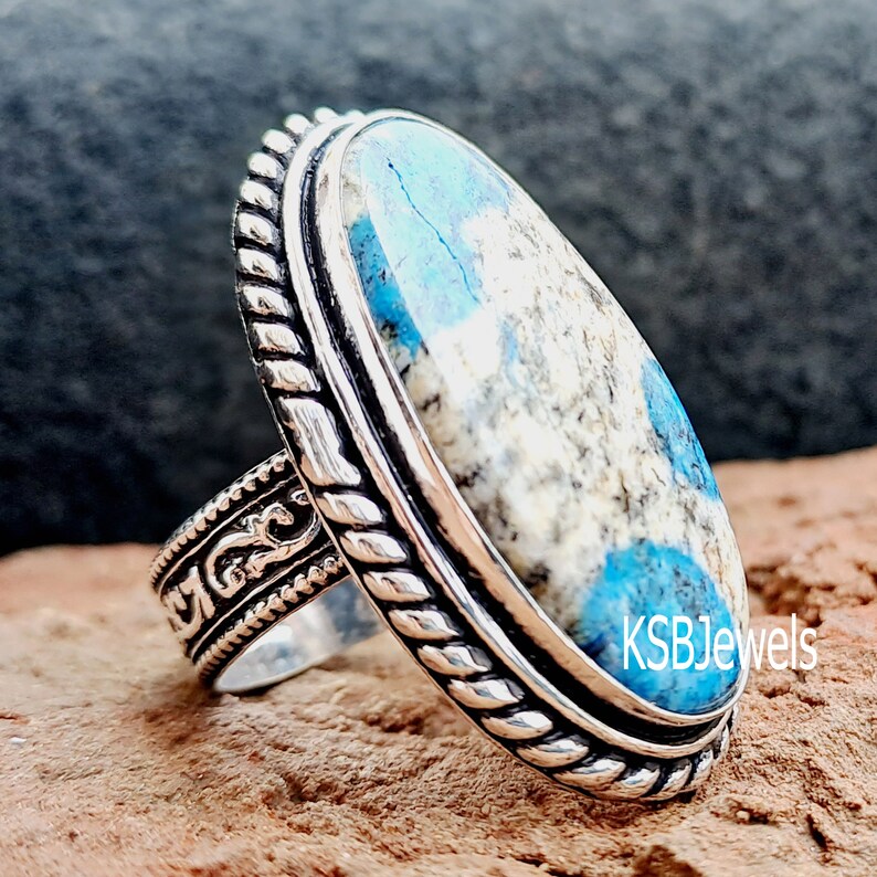 May include: A silver ring featuring a blue and white oval stone set in a rope-like design. The ring is on a brown background. KSBJewels is printed on the stone.