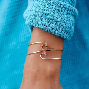 May include: Two wave-shaped bracelets on a wrist. The top bracelet is gold and the bottom bracelet is silver. The bracelets are both open and have a simple design. The text "K88Jewels" is visible on the gold bracelet.