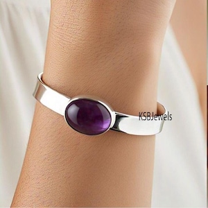Peut inclure: Un bracelet jonc en argent avec une grande pierre ovale en améthyste. Le bracelet est ouvert et a un design simple. KSBJewels est imprimé sur le bracelet.