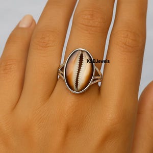 Puede incluir: Anillo ovalado con una concha natural en un engaste plateado. La concha presenta una paleta de colores cálidos, marrón y blanco. El diseño del anillo incluye bandas divididas a cada lado de la concha. El texto "KSBJewels" es visible.