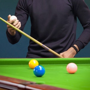 May include: A person holding a pool cue with a white cue ball on the end. The cue has the word "Preciseball" printed on it. There are three billiard balls on a green felt snooker table: a yellow ball, a blue ball, and a white ball.
