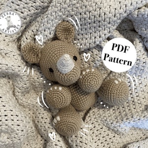 May include: A crocheted amigurumi rhinoceros toy in brown and white, lying on a white knitted blanket. The toy has a white horn and small white dots on its feet. The text "PDF Pattern" is visible in a white circle on the blanket.