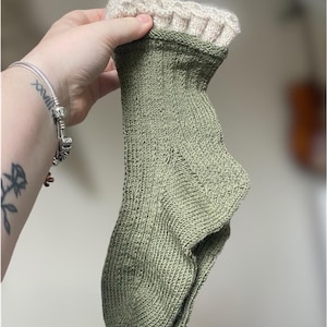 Op de afbeelding: Een groene gebreide sok met een witte gehaakte boord. De sok is dubbelgevouwen en wordt in een hand gehouden.