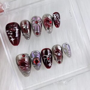 May include: A set of press-on nails in various shapes and sizes. The nails feature a color palette of burgundy, silver, and lavender. Some nails have star, bow, and pearl accents. The nails are displayed in a clear plastic container.