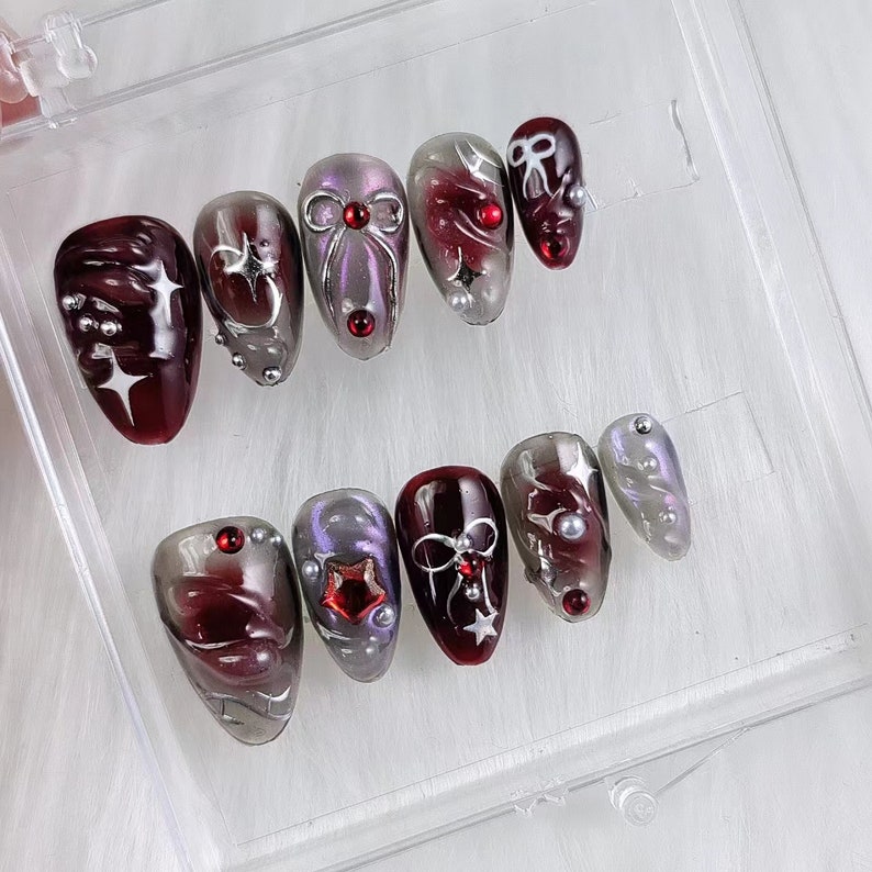 May include: A set of press-on nails in various shapes and sizes. The nails feature a color palette of burgundy, silver, and lavender. Some nails have star, bow, and pearl accents. The nails are displayed in a clear plastic container.