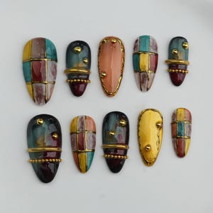 Op de afbeelding: Een verzameling van tien kunstnagels met diverse ontwerpen. De nagels tonen een mix van kleuren, waaronder teal, rood, geel en goud. Sommige nagels hebben een patchwork-ontwerp, terwijl andere effen kleuren hebben met gouden accenten. De nagels zijn gerangschikt op een wit oppervlak.