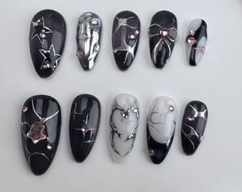 May include: Set of ten black and white press-on nails with silver and pink accents. The nails feature various designs including stars, hearts, and swirls.