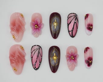 May include: A collection of ten artificial nails in various designs. Some nails feature pink and white floral accents, while others showcase butterfly wing patterns in pink and black. Additional nails have gold starburst designs and gold embellishments.