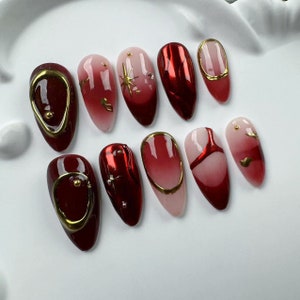 Pode incluir: Conjunto de dez unhas postiças vermelhas e douradas com vários designs. As unhas são longas e têm uma forma pontiaguda. Algumas unhas têm um efeito degradê, enquanto outras têm detalhes dourados ou um acabamento metálico.