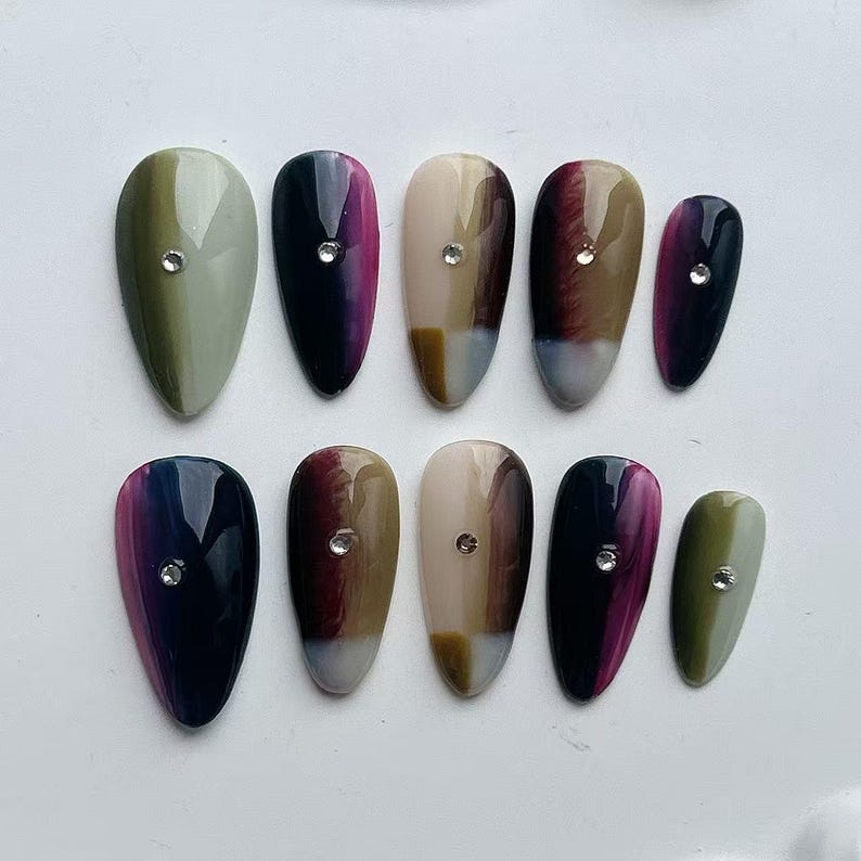 Puede incluir: Un conjunto de diez u&ntilde;as postizas ovaladas en varios colores, incluyendo verde oliva, negro, beige y burdeos, cada una con un efecto degradado y un peque&ntilde;o adorno de pedrer&iacute;a. Las u&ntilde;as est&aacute;n dispuestas sobre una superficie blanca.