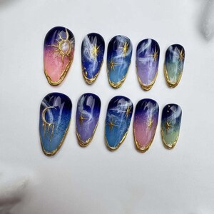 May include: Set of ten false nails with a celestial theme. The nails are painted with a gradient of blue, purple, and pink, and feature gold accents of stars, moons, and swirls.
