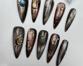 May include: Set of 11 false nails with a celestial theme. The nails are a mix of gray, brown, and black with gold accents. Each nail features a different celestial design, including stars, moons, and suns.