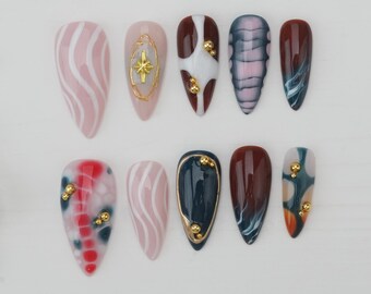 Könnte beinhalten: Eine Sammlung von zehn künstlichen Nägeln in verschiedenen Designs. Die Nägel zeigen Farben wie Rosa, Weiß, Braun, Blau und Türkis, mit goldenen Verzierungen. Einige Nägel haben Streifenmuster, andere zeigen abstrakte Kunst.