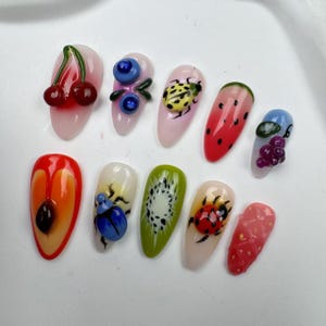 Könnte beinhalten: Zehn künstliche Nägel mit verschiedenen Designs. Die Designs beinhalten eine Kirsche, Blaubeeren, einen Marienkäfer, eine Wassermelonen-Scheibe, Trauben, eine Kiwi, einen Käfer und ein Herz.
