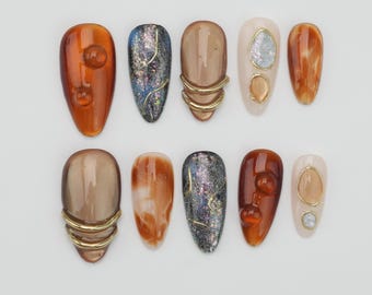 Könnte beinhalten: Eine Sammlung von zehn künstlichen Nägeln in verschiedenen Designs. Die Nägel zeigen Farben wie Bernstein, Beige und schillerndes Blau, mit goldenen Akzenten und strukturierten Oberflächen. Einige Nägel haben ein 3D-Blasendesign, andere ein Wirbelmuster.