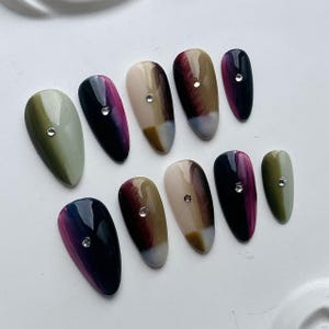 Puede incluir: Un conjunto de diez u&ntilde;as postizas ovaladas. Cada u&ntilde;a presenta un dise&ntilde;o &uacute;nico, con gradientes de color verde, morado y marr&oacute;n, y un peque&ntilde;o y brillante pedrer&iacute;a. Las u&ntilde;as est&aacute;n dispuestas sobre una superficie blanca.