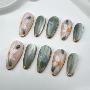 May include: Set of ten false nails with a floral design. The nails are oval shaped and have a light pink base with white flowers and green leaves. Some nails have a light green base with a silver shimmer finish.