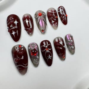 May include: A set of ten artificial nails with a dark red and silver design. Each nail features a unique pattern, including silver embellishments, star shapes, and red accents. The nails have an oval shape and a glossy finish.