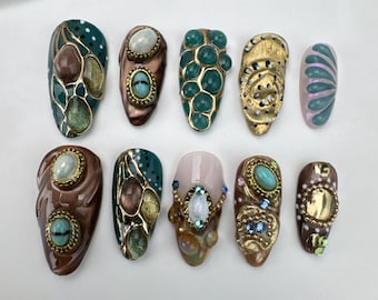 May include: A set of ten artificial nails with intricate designs. The nails feature a variety of colors, including teal, gold, brown, and pink, and are adorned with gemstones and detailed patterns. Each nail has a unique design.