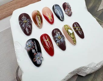 Può includere: Set di dieci unghie finte con vari design. Le unghie sono a forma di mandorla e presentano accenti rossi, neri e dorati. Alcune unghie hanno delicati disegni floreali, mentre altre hanno linee dorate e glitter.